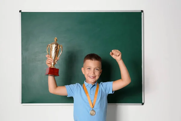 Gelukkige jongen met gouden beker en medaille in de buurt van schoolbord in de klas — Stockfoto