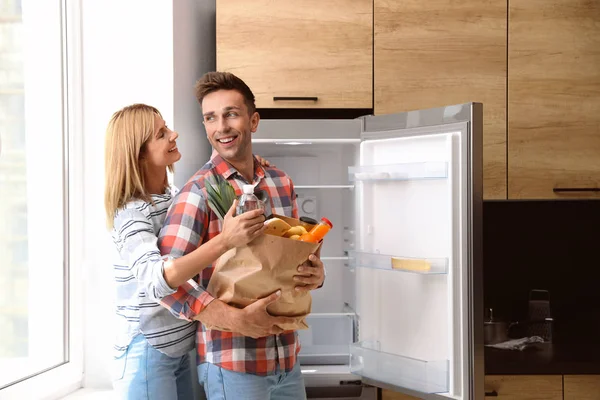Ευτυχισμένο ζευγάρι με χάρτινη σακούλα γεμάτη προϊόντα κοντά στο ψυγείο στην κουζίνα — Φωτογραφία Αρχείου