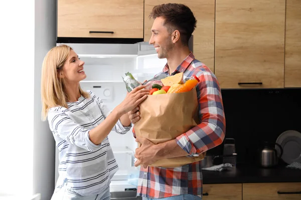 Ευτυχισμένο ζευγάρι με χάρτινη σακούλα γεμάτη προϊόντα κοντά στο ψυγείο στην κουζίνα — Φωτογραφία Αρχείου
