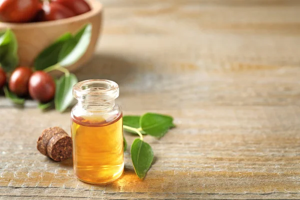 Glass bottle with jojoba oil and leaves on wooden table. Space for text