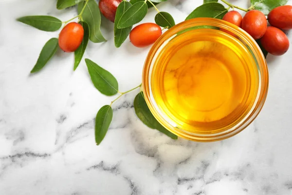 Glass bowl with jojoba oil and seeds on white marble table, flat lay — Stock Photo, Image