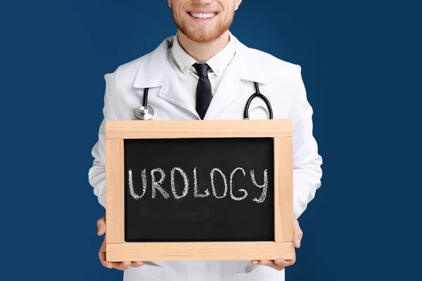 Homme médecin tenant petit tableau noir avec mot UROLOGIE sur fond bleu, gros plan — Photo