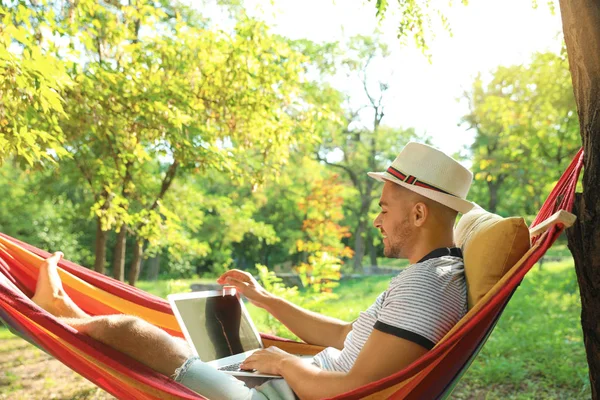 Jonge man met laptop rusten in comfortabele hangmat in groene tuin — Stockfoto