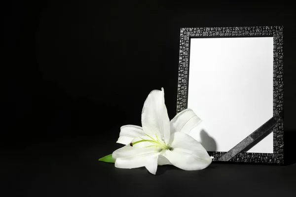 Funeral photo frame with ribbon and white lily on dark table against black background. Space for design — ストック写真