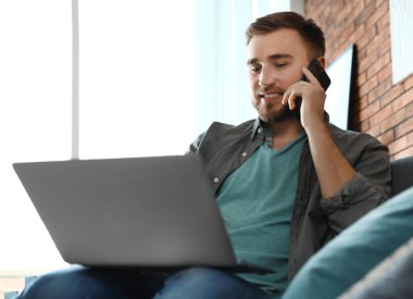 Oturma odasında dizüstü bilgisayar kullanırken telefonda konuşan genç adam