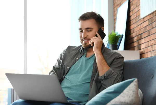 Młody człowiek rozmawia na telefon podczas korzystania z laptopa w salonie — Zdjęcie stockowe