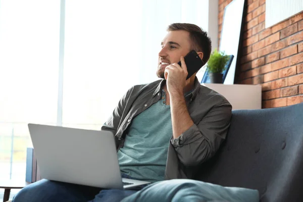 Hombre joven hablando por teléfono mientras usa el ordenador portátil en la sala de estar —  Fotos de Stock