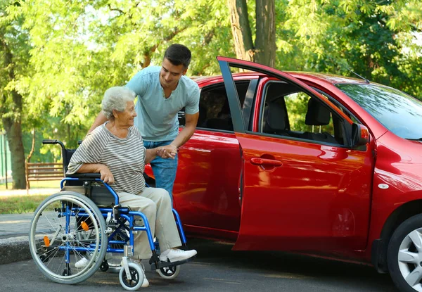 Ung man hjälper handikappade Senior Kvinna i rullstol att komma in i bilen utomhus — Stockfoto