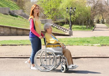 Senior woman in wheelchair with young assistant at park on sunny day