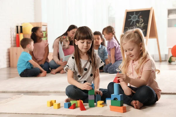 Милые девочки играют со строительными блоками на полу, в то время как воспитатель детского сада читает книги другим детям в помещении — стоковое фото