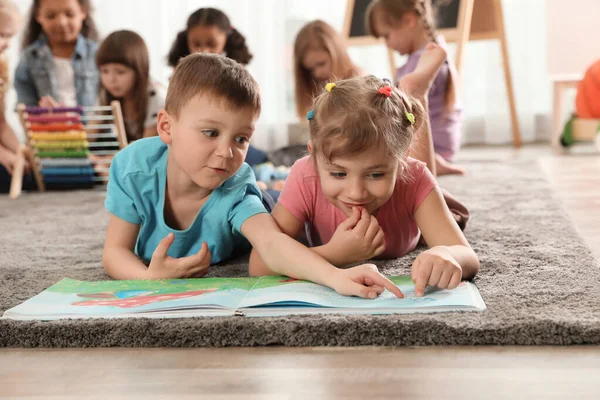 Roztomilé děti čtení knihy na podlaze, zatímco ostatní děti hrají spolu ve školce — Stock fotografie