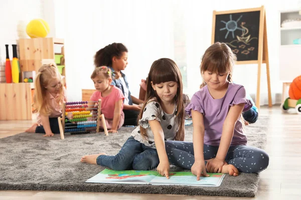Roztomilé dívky čtení knihy na podlaze, zatímco ostatní děti spolu hrají ve školce — Stock fotografie