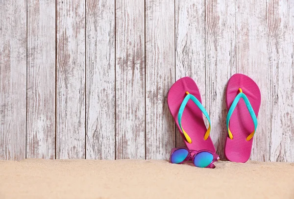 Infradito e occhiali da sole luminosi sulla sabbia vicino alla parete di legno, spazio per il testo. Accessori spiaggia estate — Foto Stock