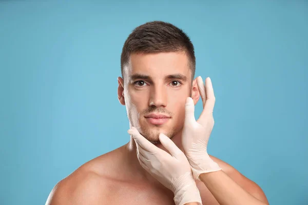 Médico examinando o rosto do homem para cirurgia estética em fundo azul claro — Fotografia de Stock