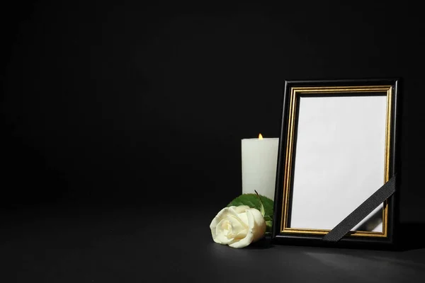 Moldura de foto fúnebre com fita, rosa branca e vela na mesa escura contra fundo preto. Espaço para design — Fotografia de Stock