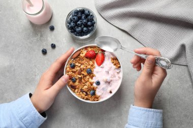 Woman eating tasty homemade granola with yogurt and berries at grey table, top view. Healthy breakfast