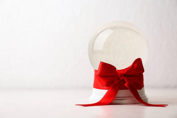 Piękna świąteczna kula śnieżna z czerwoną kokardą na stole na jasnym tle, miejsce na tekst — Zdjęcie stockowe