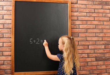 Cute little left-handed girl doing sums on chalkboard near brick wall clipart
