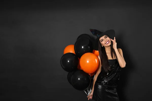 Mulher bonita vestindo traje de bruxa com balões para festa de Halloween em fundo preto, espaço para texto — Fotografia de Stock
