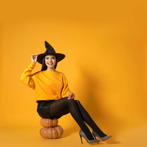 Hermosa mujer con traje de bruja para la fiesta de Halloween sentado en calabazas contra el fondo amarillo, espacio para el texto —  Fotos de Stock