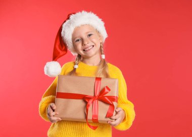 Kırmızı arka planda hediye kutusu olan Noel Baba şapkalı küçük mutlu çocuk. Noel kutlaması