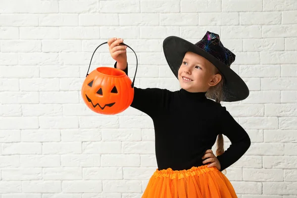 Carino bambina con zucca secchio di caramelle indossando costume di Halloween vicino muro di mattoni bianchi — Foto Stock