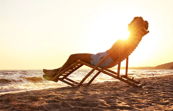 Jovem relaxante na cadeira deck na praia perto do mar — Fotografia de Stock