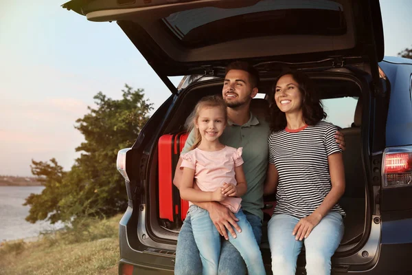 Счастливая молодая семья сидит в багажнике машины на берегу реки — стоковое фото