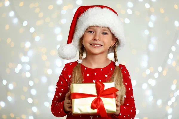サンタの帽子の幸せな小さな子供は、ぼやけたお祝いの光に対してギフトボックスを持ちます。クリスマスのお祝い — ストック写真