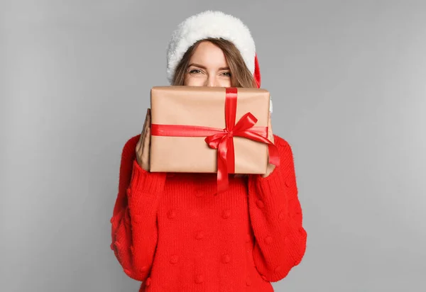 Jovem feliz em chapéu de Papai Noel e suéter com caixa de presente em fundo cinza claro. Celebração de Natal — Fotografia de Stock