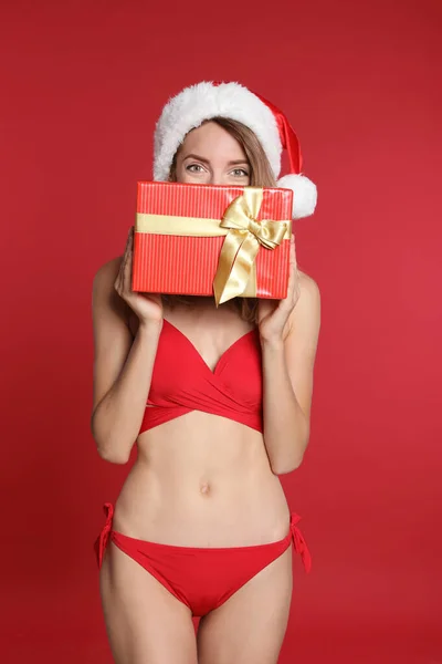 Kırmızı arka planda hediye kutusu olan bikinili ve Noel Baba şapkalı seksi genç kadın. Noel kutlaması — Stok fotoğraf