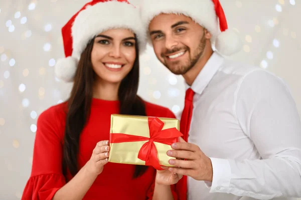 Счастливая молодая пара в шляпах Санты с подарочной коробкой против размытых праздничных огней. Праздник Рождества — стоковое фото