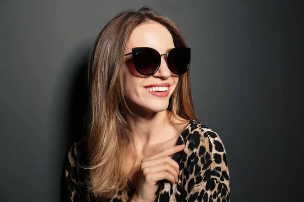 Jeune femme portant des lunettes de soleil élégantes sur fond gris foncé — Photo