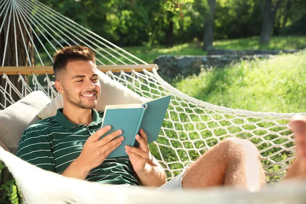 Jonge man lezen boek in comfortabele hangmat op groene tuin — Stockfoto