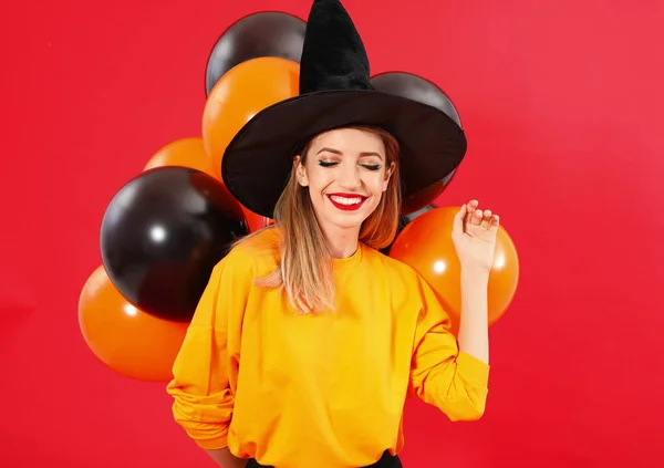 Schöne Frau trägt Hexenkostüm mit Luftballons für Halloween-Party auf rotem Hintergrund — Stockfoto