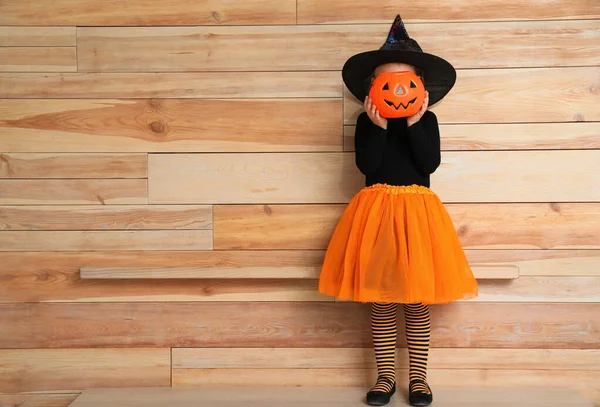 Menina bonito com balde de doces de abóbora vestindo traje de Halloween perto da parede de madeira. Espaço para texto — Fotografia de Stock