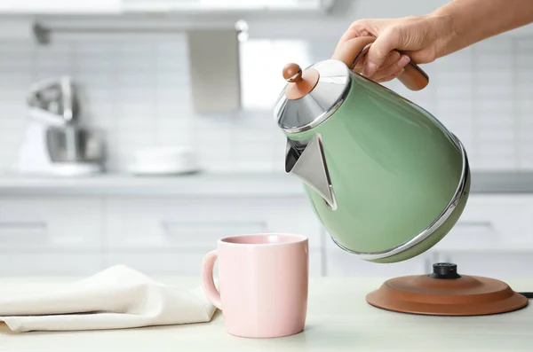 Vrouw gieten water uit moderne elektrische waterkoker in beker aan houten tafel in de keuken, close-up — Stockfoto