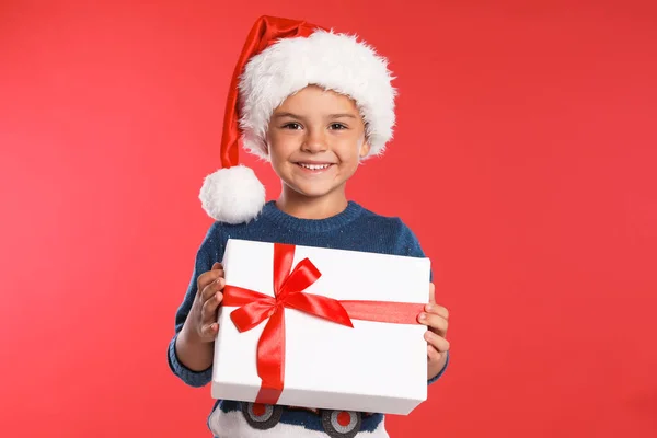 Criança feliz em chapéu de Papai Noel com caixa de presente no fundo vermelho. Celebração de Natal — Fotografia de Stock
