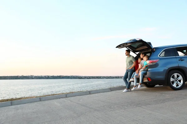 Feliz joven familia sentada en el maletero del coche a orillas del río — Foto de Stock