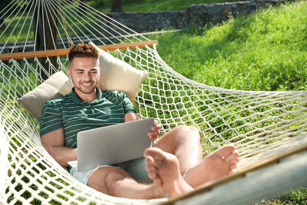 Junger Mann mit Laptop ruht in bequemer Hängematte im grünen Garten — Stockfoto