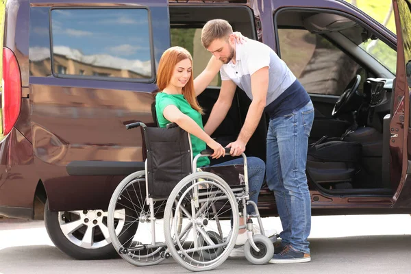 Młody mężczyzna pomaga kobiecie usiąść na wózku inwalidzkim w pobliżu vana na zewnątrz — Zdjęcie stockowe