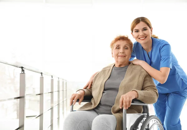 Νοσοκόμα βοηθώντας ηλικιωμένη γυναίκα σε αναπηρικό αμαξίδιο σε εσωτερικούς χώρους. Χώρος για κείμενο — Φωτογραφία Αρχείου