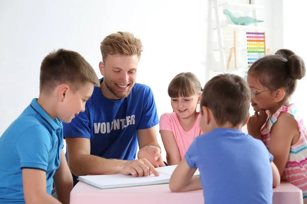 Young volunteer reading book with children at table indoors — Stock Photo, Image