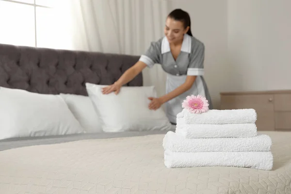 Stack of clean towels with flower on bed and blurred chambermaid in hotel room, closeup. Space for text — Stock Photo, Image