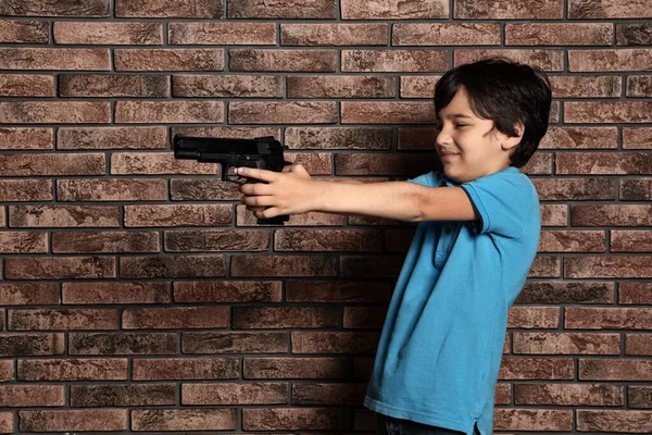 Un niño jugando con un arma contra una pared de ladrillo. Peligroso juego — Foto de Stock