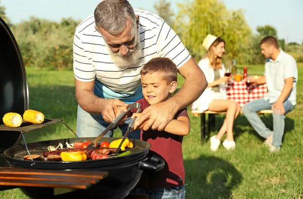 Bunicul cu un băiețel gătit mâncare pe grătar și familia lor în parc — Fotografie, imagine de stoc