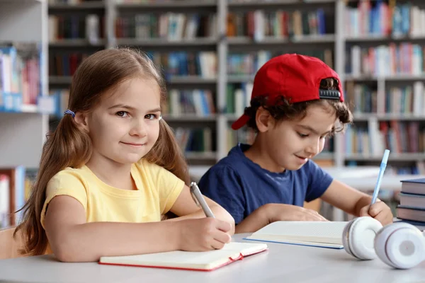 Crianças escrevendo à mesa com livros na sala de leitura da biblioteca — Fotografia de Stock