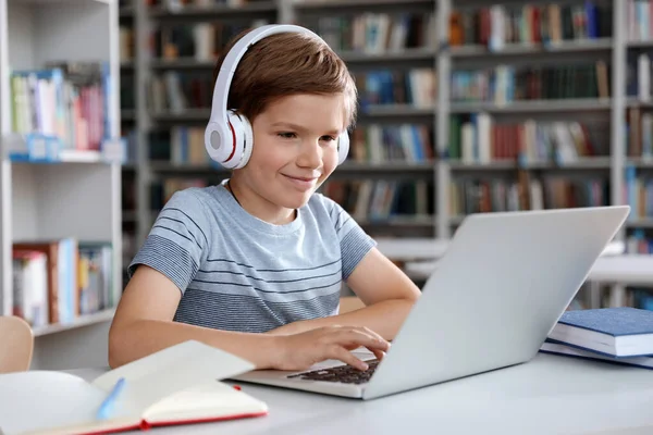 Malý chlapec se sluchátky čtení knihy pomocí notebooku v knihovně — Stock fotografie
