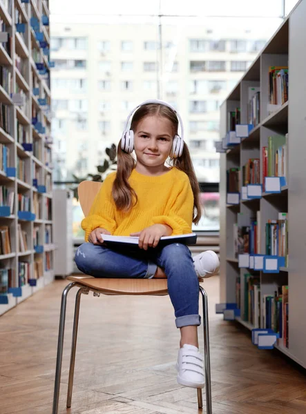 Linda niña con auriculares lectura libro en silla en la biblioteca — Foto de Stock