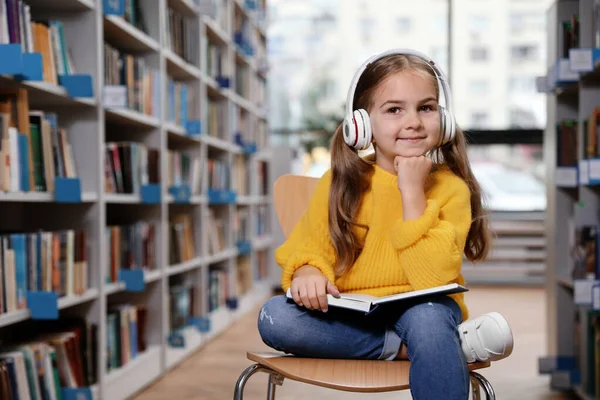 Linda niña con auriculares lectura libro en silla en la biblioteca — Foto de Stock